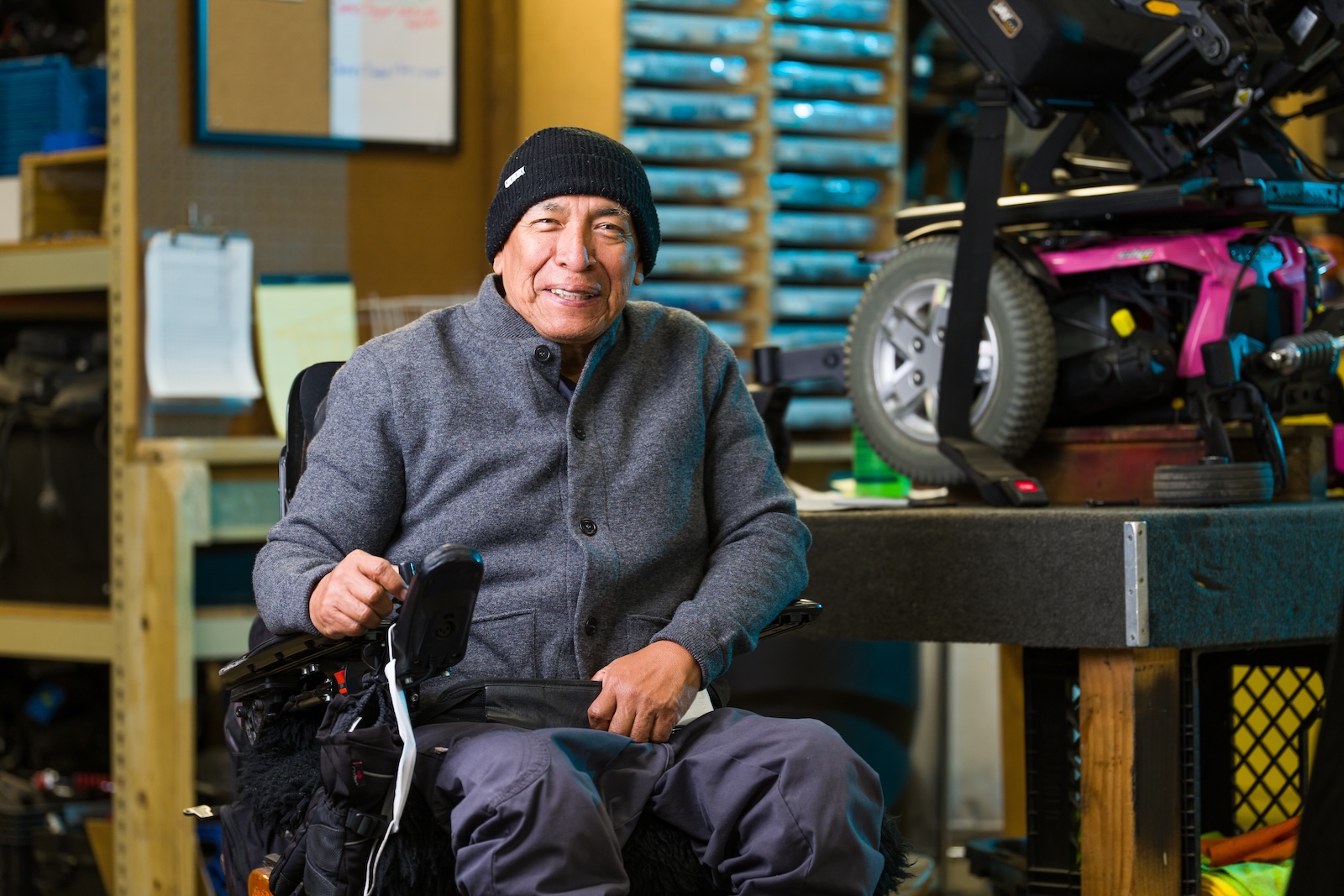 Man in wheelchair sits next to a wheelchair in repair in a shop.
