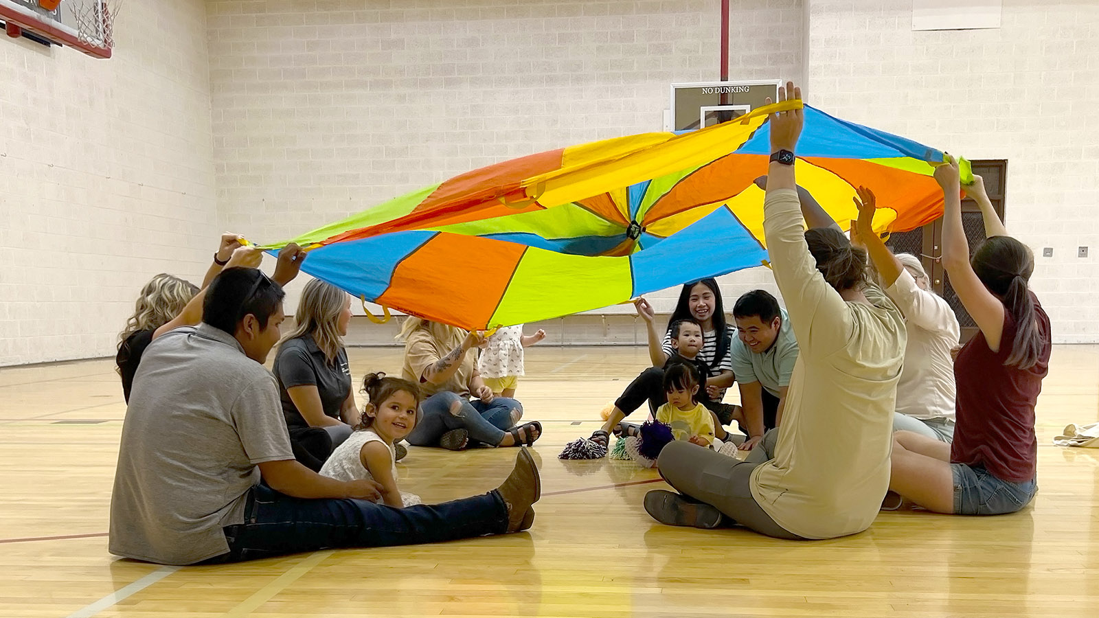 a group of parents and children plays with a parachute in a gym