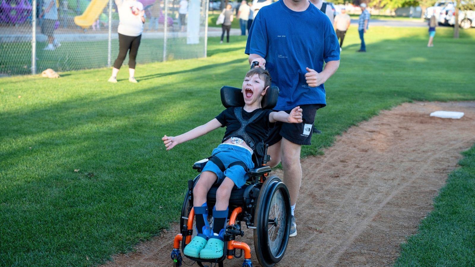 A young boy in a wheelchair is pushed towards home plate on a baseball field. His face is fully of joy.