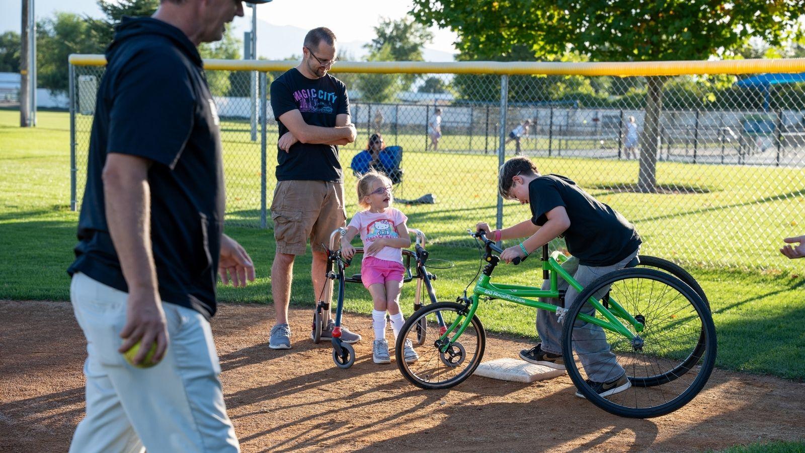 Individuals at Top Sports Teeball stand at 1st plate. One individual uses a frame runner. Another individual uses a walker with wheels.