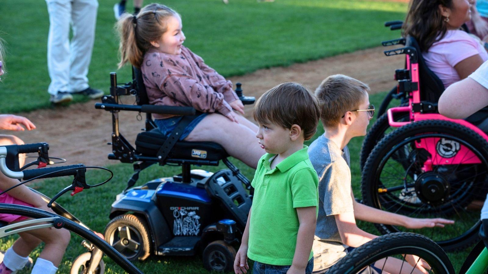 A group of individuals gather by the dugout at a baseball field. Some individuals are using assistive technology such as power wheelchairs.
