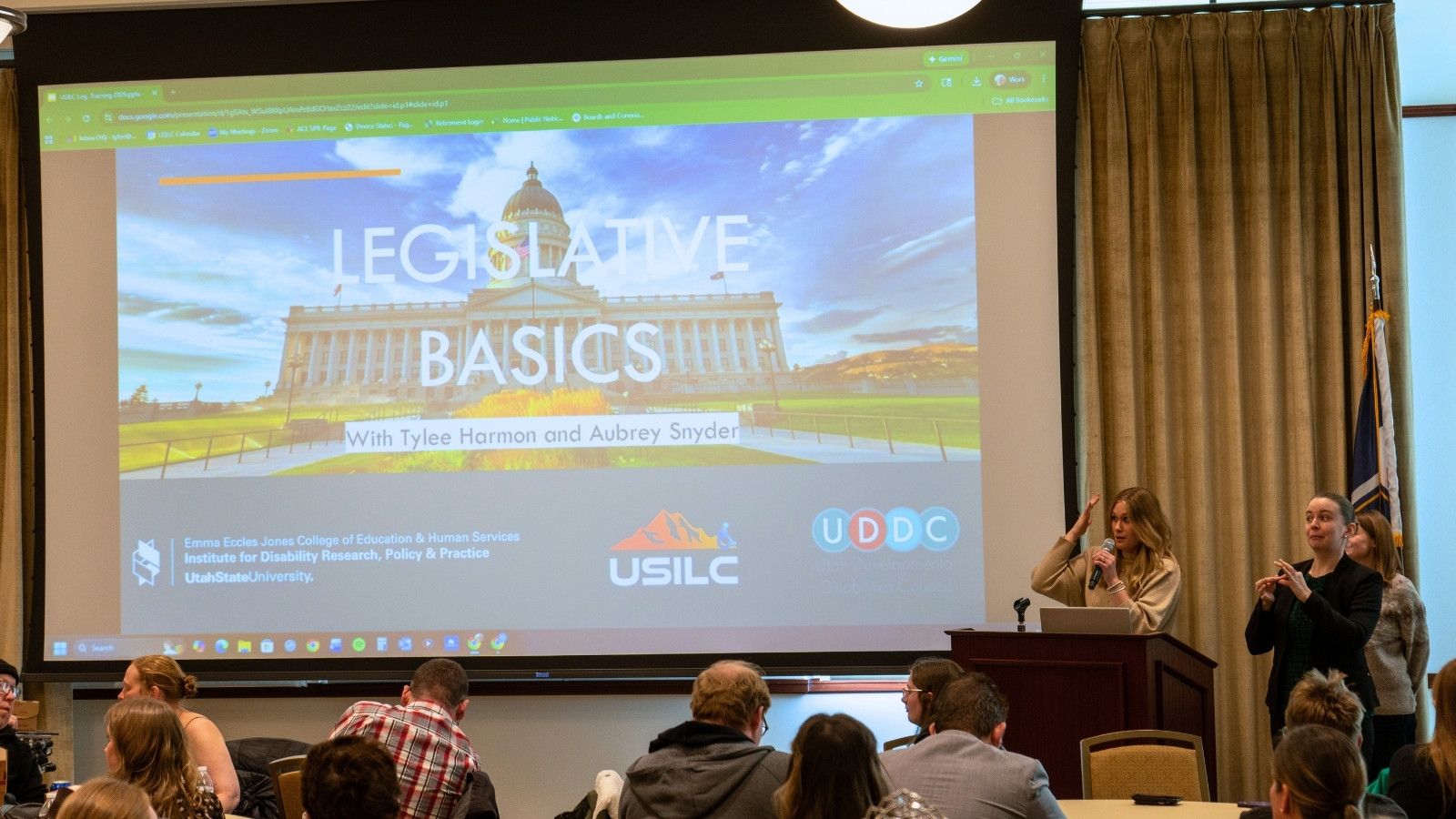 Presenters speak to a crowd of people with a presentation on the screen titled: Legislative Basics.