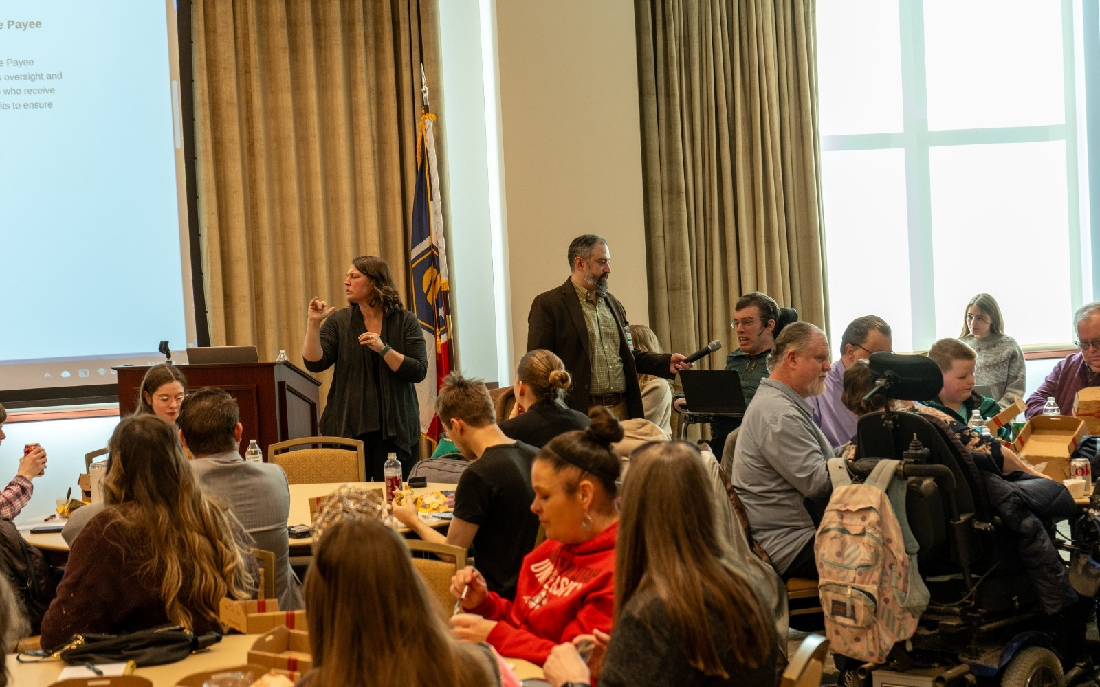 A room of people at tables. A man holds a mic up to an individual in a wheelchair as they speak.